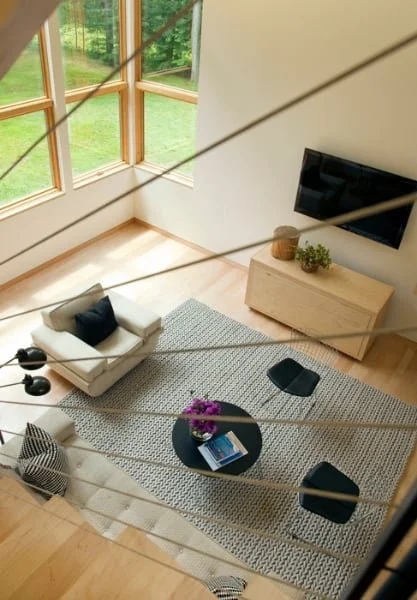 A modern living room with large windows, a white armchair with black pillows, a gray area rug, a round coffee table with purple flowers and books, a black TV mounted on the wall, and a wooden TV stand with decorative items.