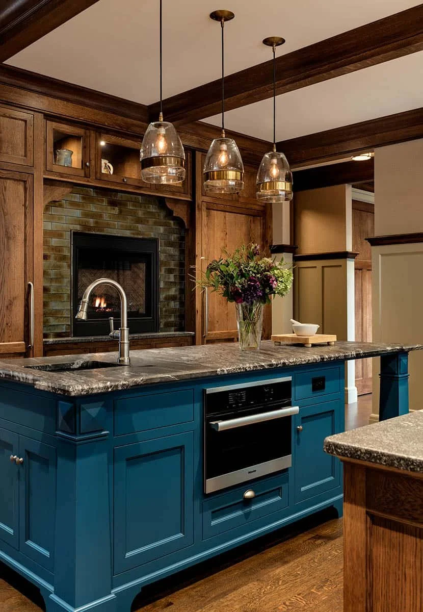 A kitchen with a blue island, marble countertop, stainless steel oven, a faucet, hanging glass pendant lights, a flower arrangement, and a brick fireplace in the background.