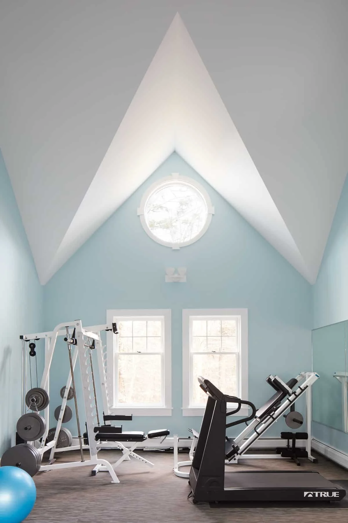 Home gym with light blue walls, two windows, a round window near the ceiling, and exercise equipment including a treadmill, weight machines, and a blue exercise ball.