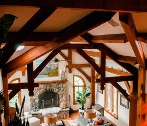 Interior view of a cozy living room with wooden beams, a stone fireplace, large windows, and indoor plants.