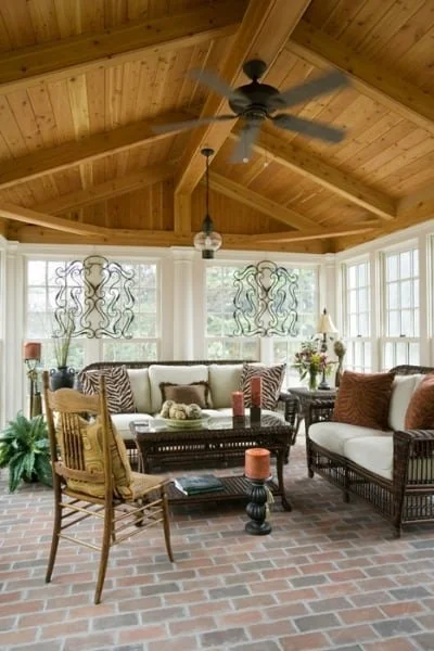 Sunroom with a wooden vaulted ceiling, a ceiling fan, wicker and wooden furniture, and large windows with decorative curtains.
