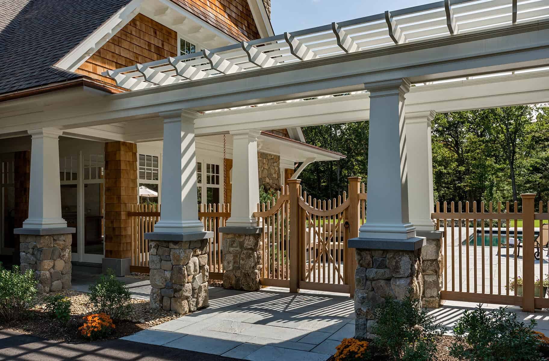 A house with a porch supported by white columns with stone bases, a wooden gate, and a fenced backyard with a swimming pool, surrounded by trees and landscaping.