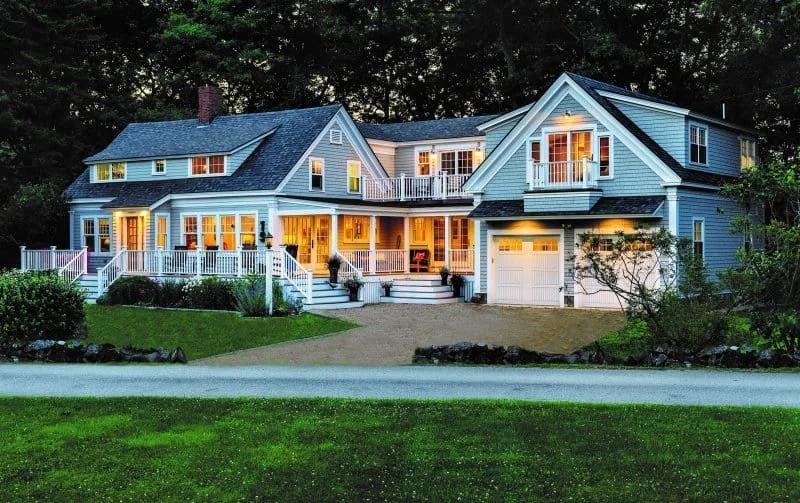 Large two-story house with a white exterior, front porch, multiple windows, and a garage, illuminated at dusk, surrounded by green lawn and trees.