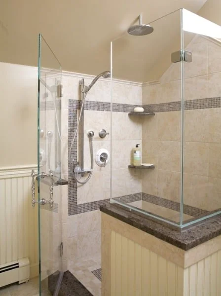 A walk-in shower with glass panels, a rain shower head, a handheld shower, and built-in shelves with soap and shampoo bottles in a tiled bathroom.