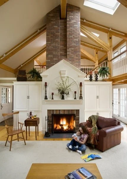 Living room with a corner fireplace, brick chimney, and vaulted ceilings with exposed wooden beams. A woman and child sit near the fireplace, with a brown sofa and a wooden chair nearby. Large windows and a skylight provide natural light.