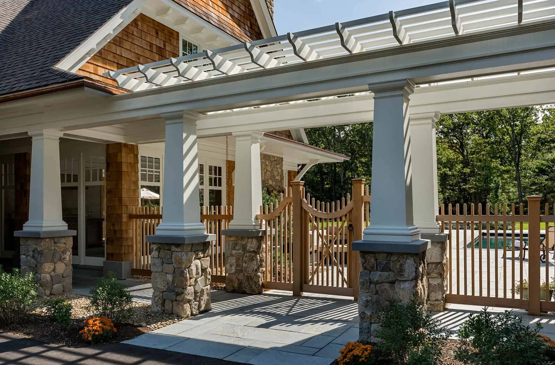 A house with a stone foundation and wooden siding, featuring a porch with white columns and a pergola overhead. There is a wooden gate and a backyard with a pool and green trees in the background.