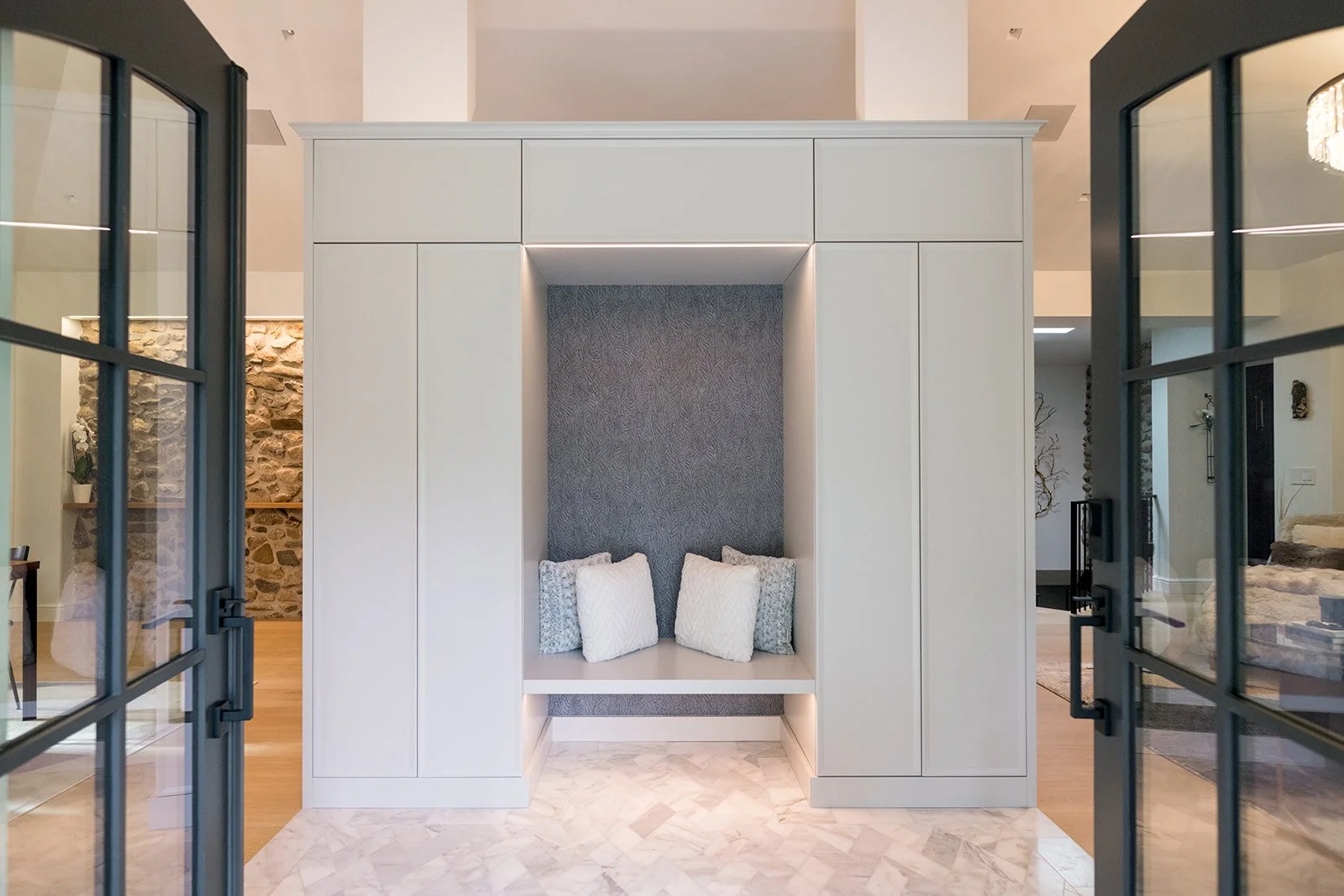 Modern built-in seating nook with gray textured wall and four white and gray pillows, framed by white cabinetry, with glass-paned doors on either side leading into a living space.