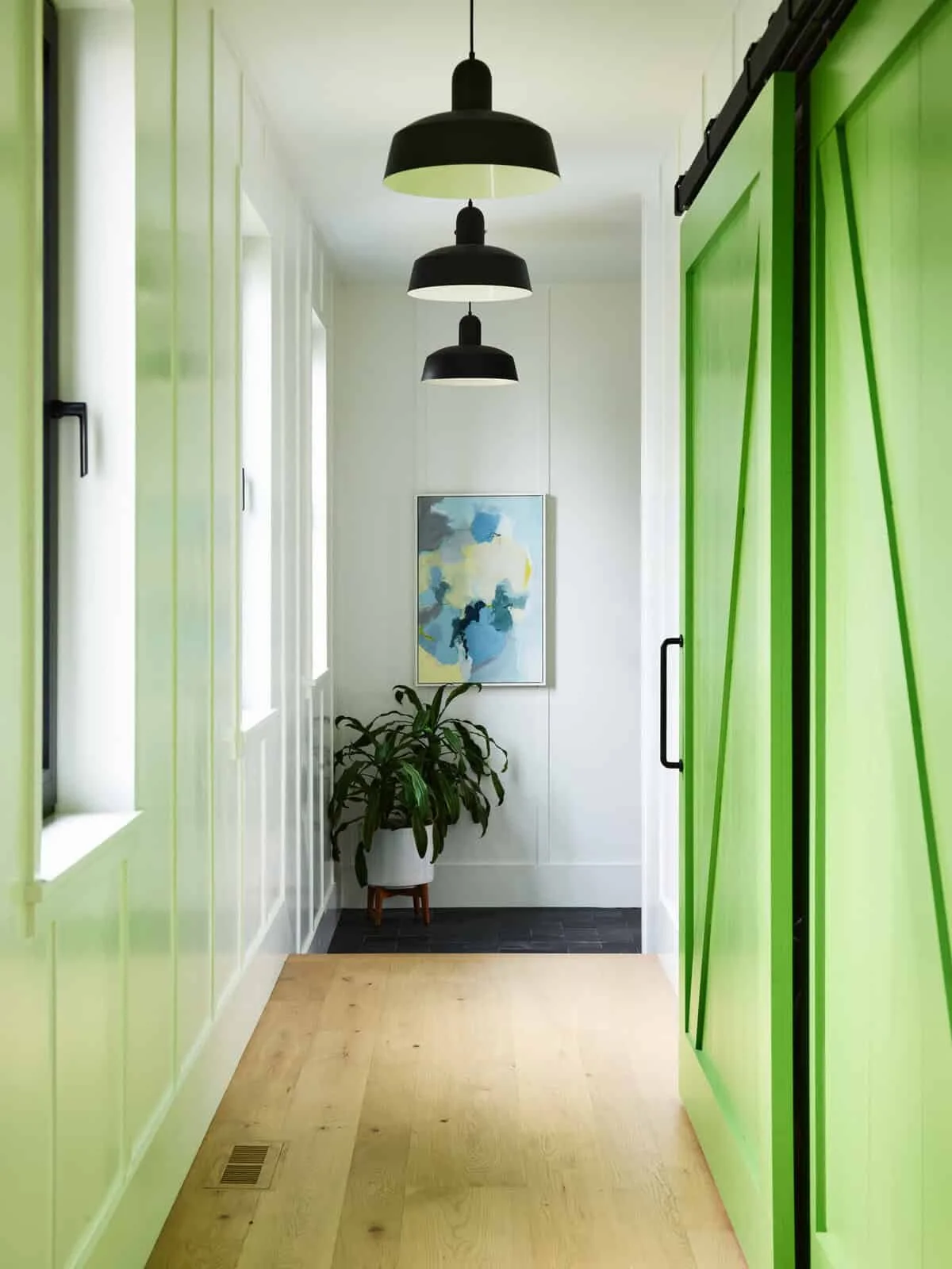 Bright hallway with lime green sliding doors on the right and matching panels on the left, black pendant lights hanging from the ceiling, a potted plant, and a blue abstract painting on the wall.