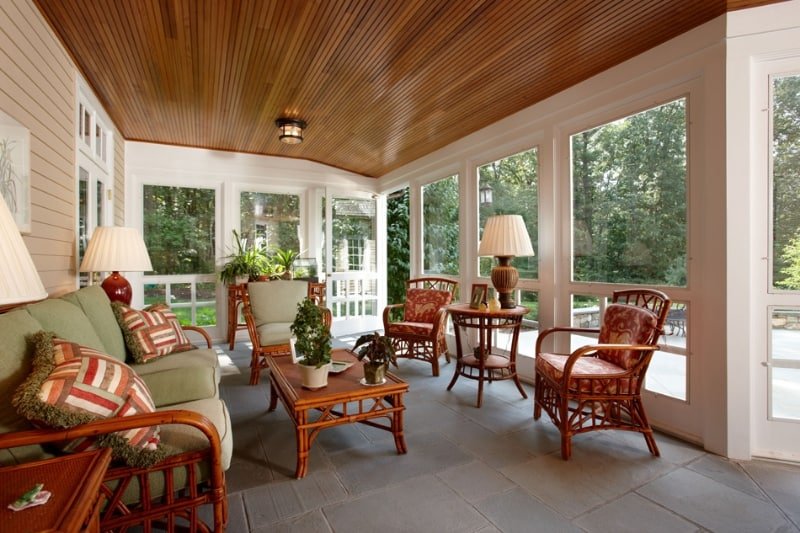 Sunroom with wicker chairs, cushioned sofa, wooden coffee table, lamps, and large windows showing greenery outside.