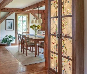 View of a dining area with a wooden table, chairs, a pendant light, and a bouquet of yellow flowers in a vase, with windows showing natural light, and a decorative door with colorful glass panels.