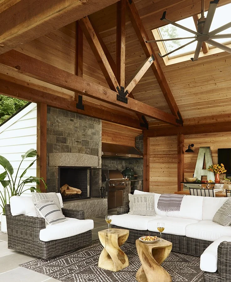 Living room with white wicker sectional sofa, wooden coffee tables, fireplace, potted plant, and wood-paneled ceiling with exposed beams and skylight.