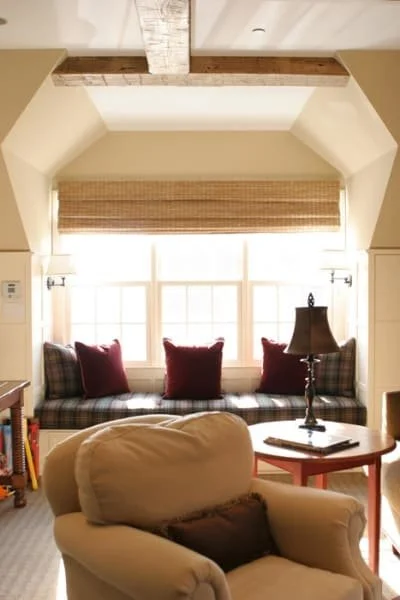 Living room with large window seat and three pillows, window with blinds, ceiling with exposed wood beam, reading lamp on side table, beige armchair in foreground