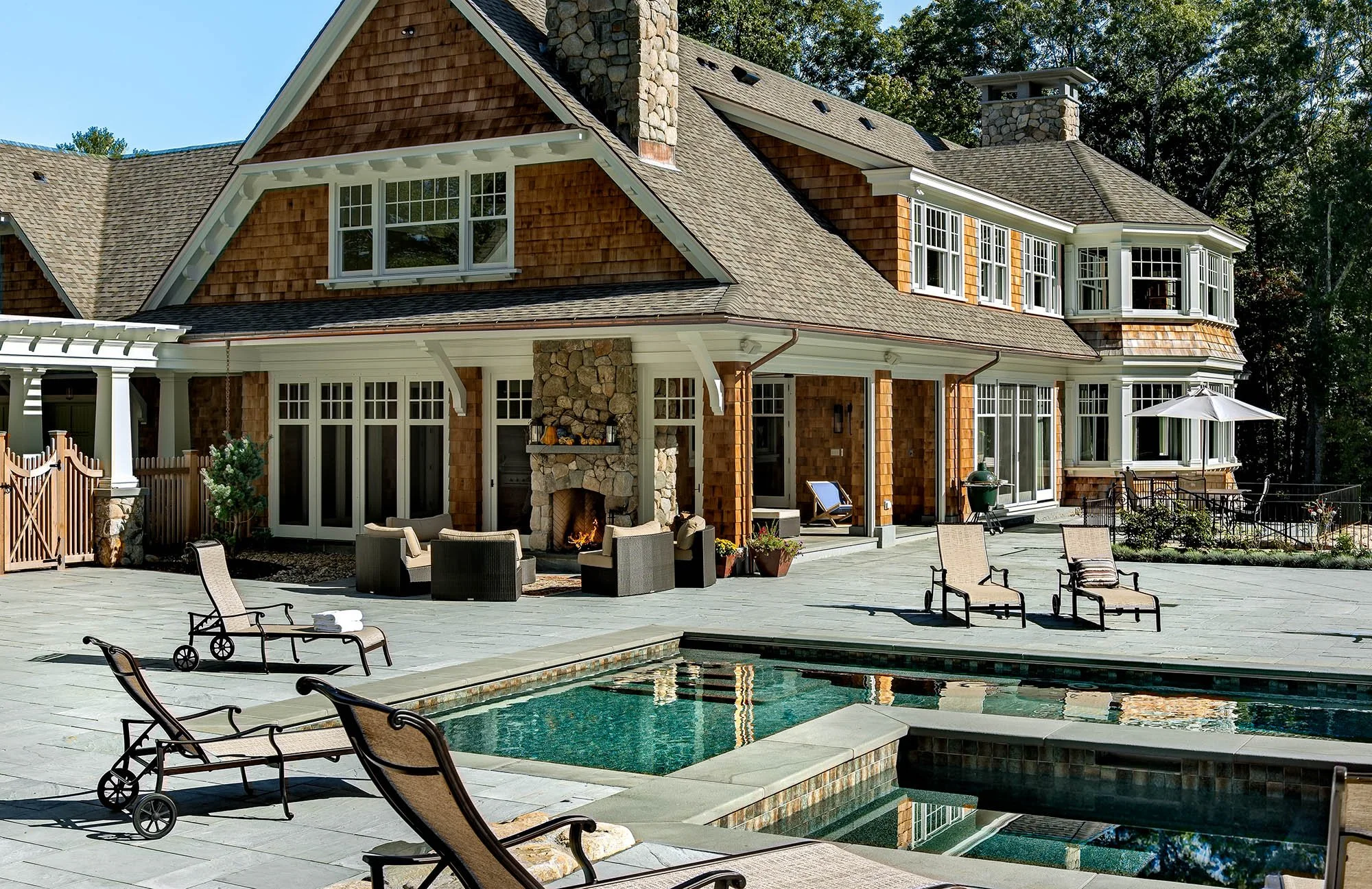 Backyard of a large house featuring a swimming pool with lounge chairs, a stone fireplace, and patio furniture, surrounded by trees.