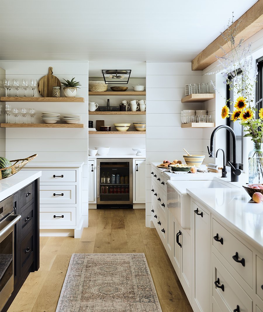 A bright farmhouse-style kitchen with white cabinets and open wooden shelves, a window with sunflowers, and a wooden countertop.