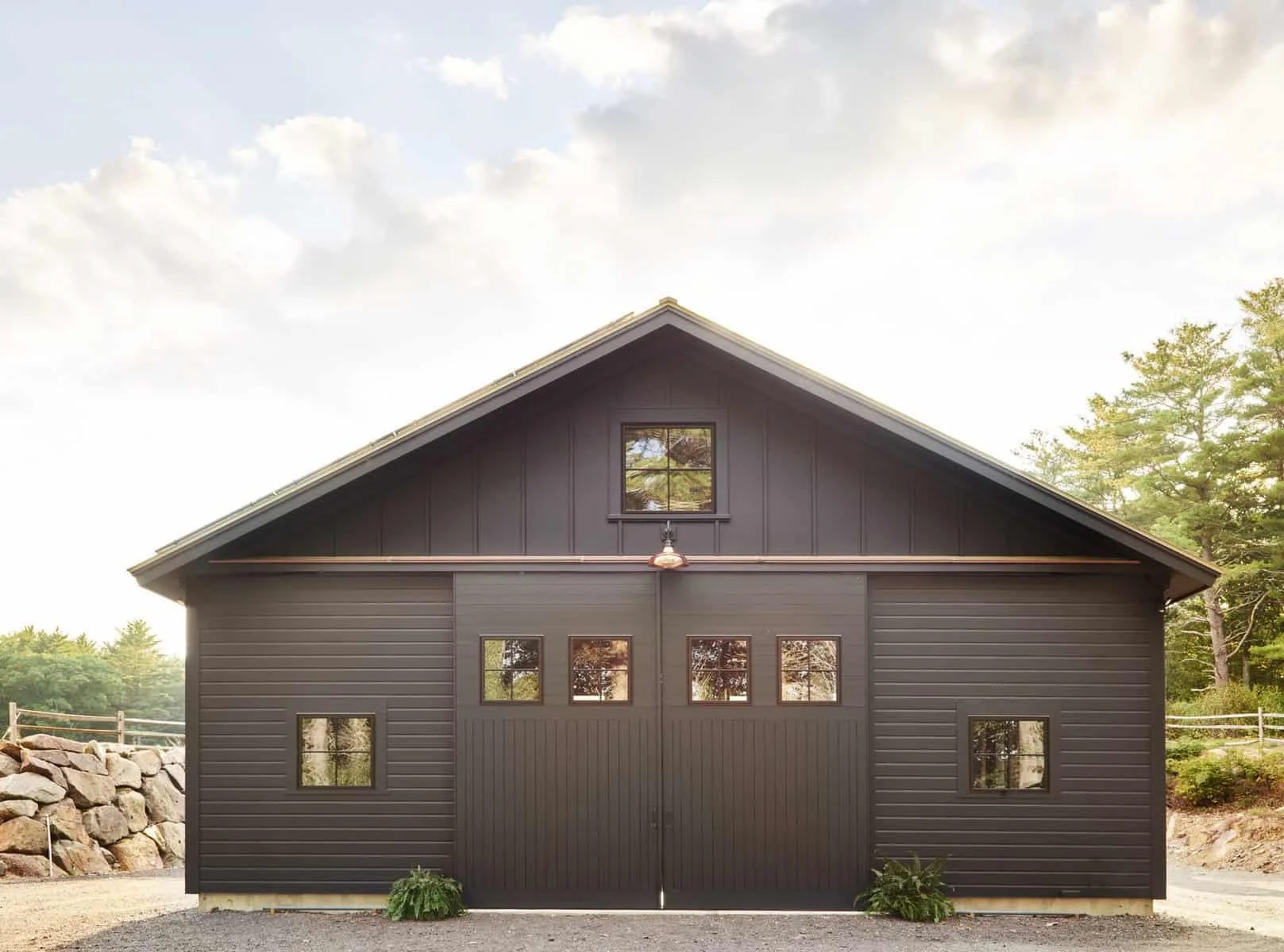 Front view of a black barn-style building with a peaked roof and six small windows on the lower level. A small window is centered on the upper level with a tree reflection visible. The building is surrounded by green trees and a stone wall on the lef