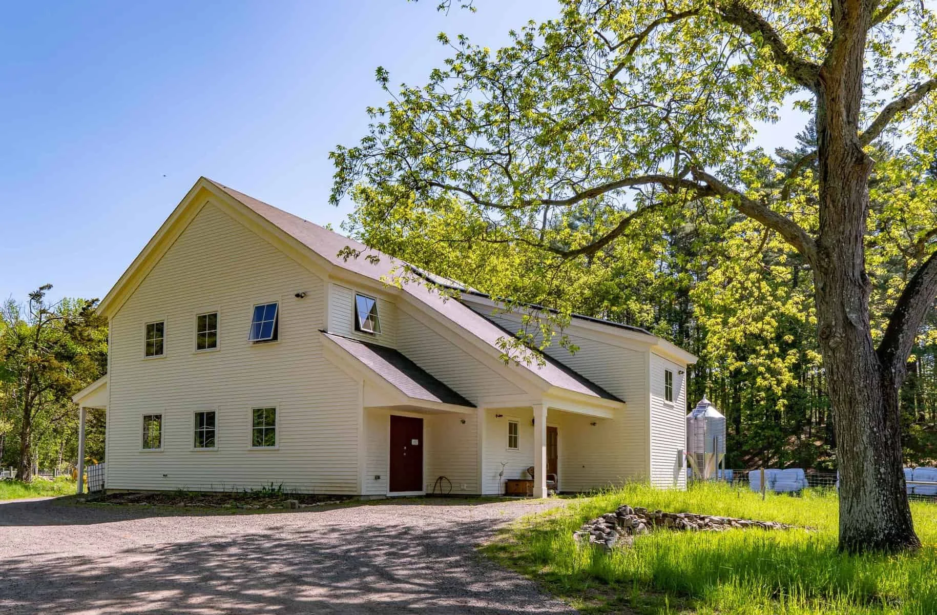 White two-story house with a sloped roof surrounded by green trees, a gravel driveway, and a grassy yard.