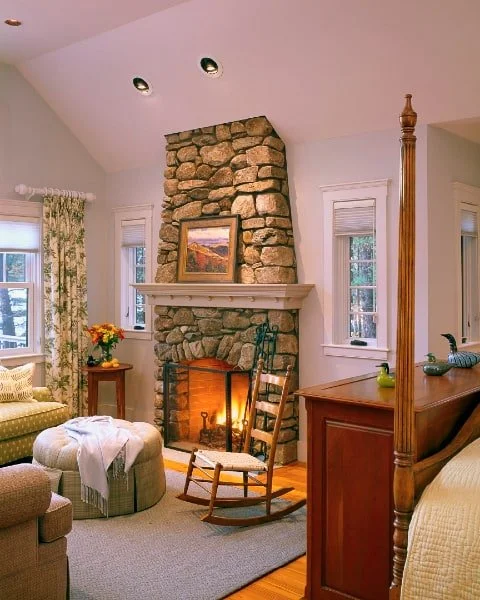 Cozy living room with a stone fireplace, two windows with floral curtains, a rocking chair in front of the fireplace, a small side table with a flower arrangement, and part of a wooden bed with a yellow blanket visible.