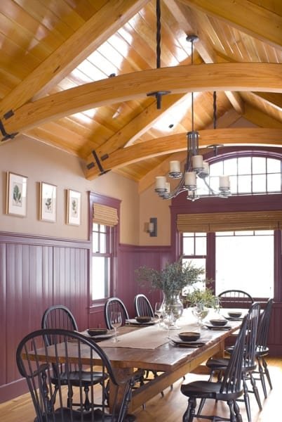 Dining room with wooden beams on the ceiling, purple wainscoting, and a large window with blinds, set with a wooden table and black chairs, decorated with a vase of flowers.