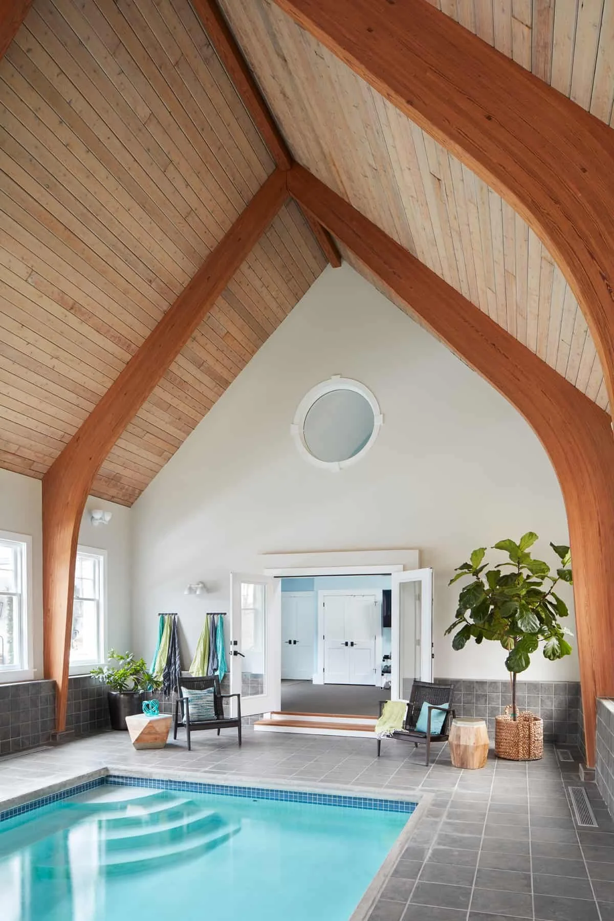 Indoor pool area with a vaulted wooden ceiling, white walls, and large windows. There is a sitting area with chairs, a potted plant, and towels hanging on hooks. The pool has clear water and is surrounded by gray tile flooring.
