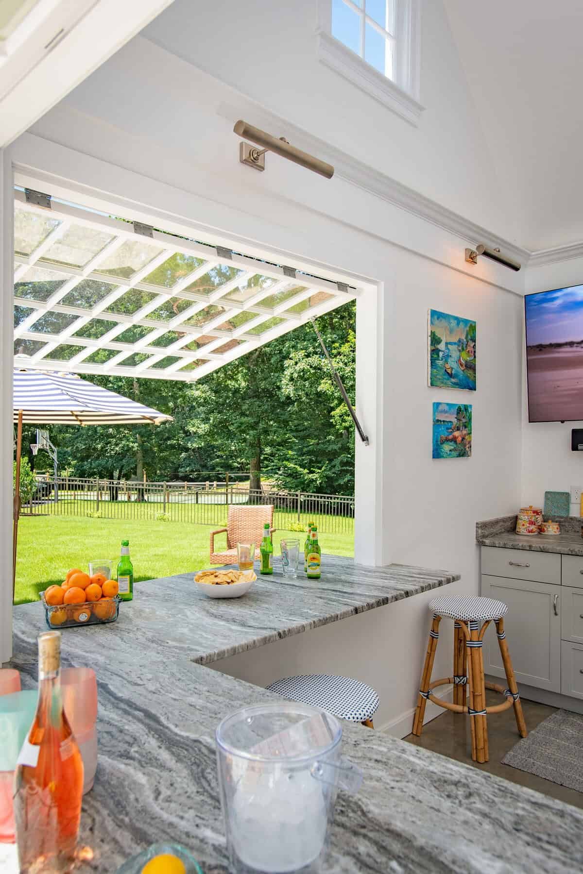 View of a bright kitchen with a view outside showing a backyard with a fence, green grass, and trees. The kitchen has a marble counter with drinks, a bowl of chips, oranges, and bottles of soda. There are two bar stools, and the wall has colorful pai
