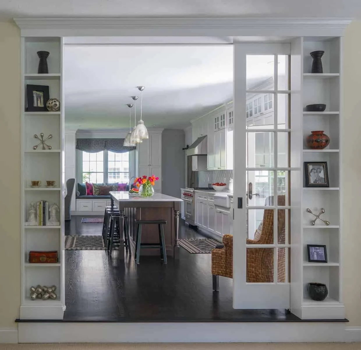 View through a white framed doorway into a bright kitchen with dark hardwood floors, white cabinets, a large island with black stools, and a cozy window seat with pink and purple throw pillows. A center vase with colorful flowers is on the island, an