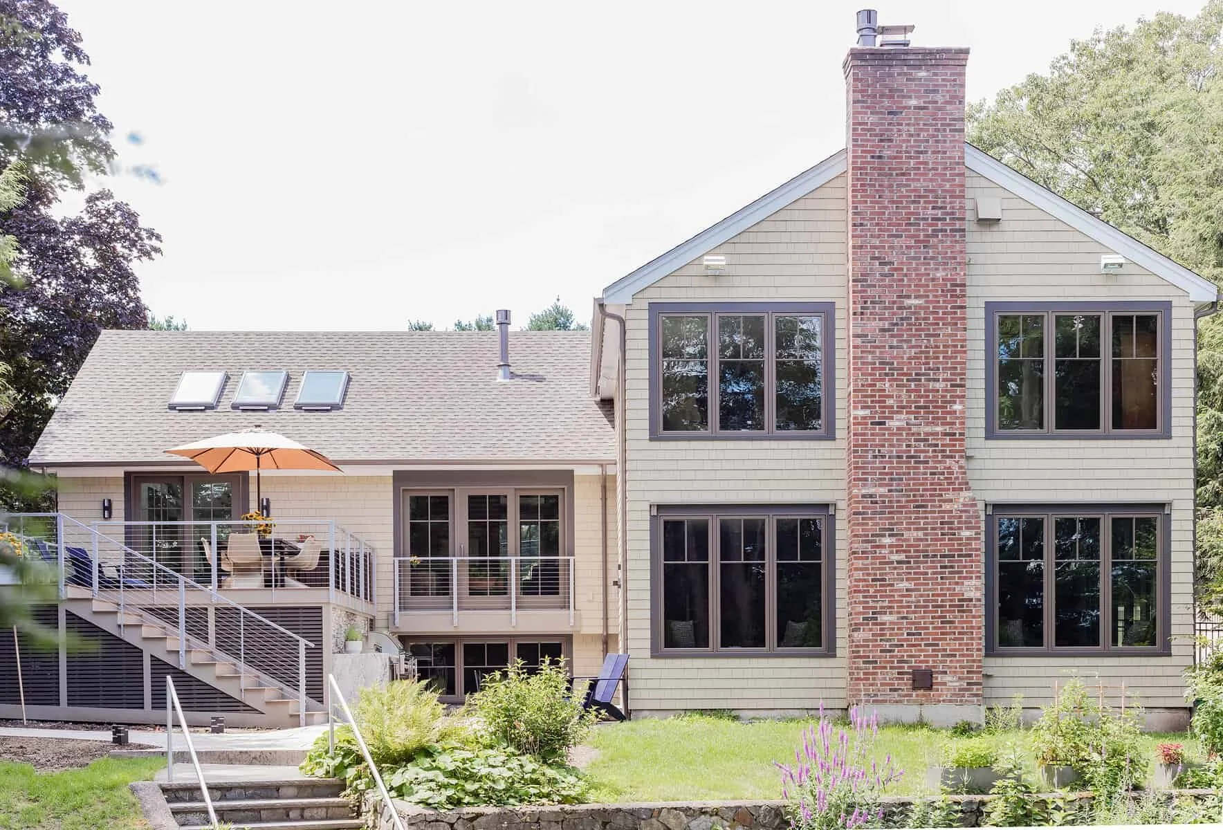 Back view of a two-story house with a brick chimney, large windows, and a balcony with outdoor furniture and an umbrella, surrounded by trees and a garden.