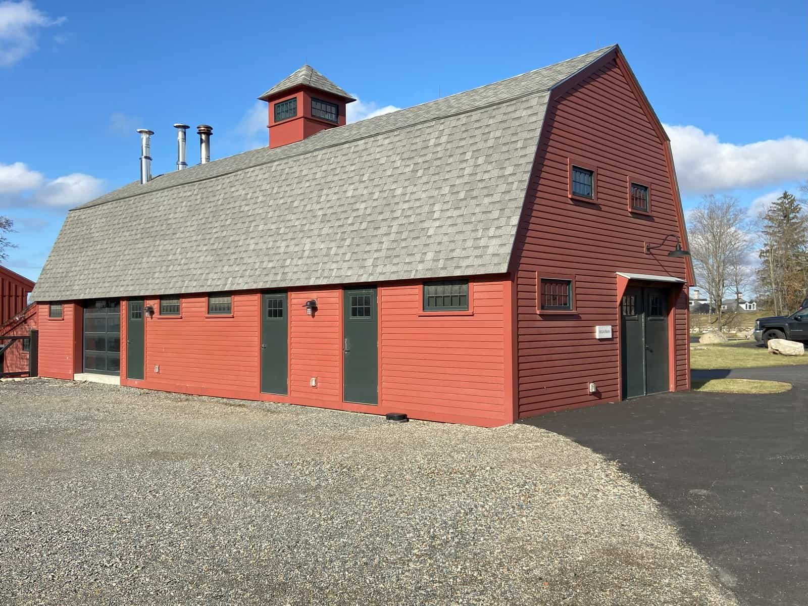 A large red barn with a gray shingled roof, six windows, and six doors, located on a gravel and asphalt lot with trees and other buildings in the background under a blue sky with clouds.