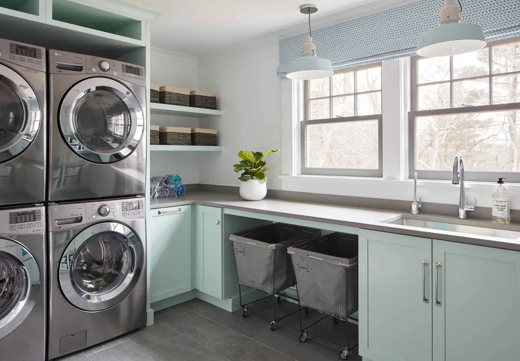 Laundry room with stacked metallic washers and dryers, light blue cabinets, white countertop, three windows, hanging light fixtures, potted plant, folded towels, and laundry hampers.