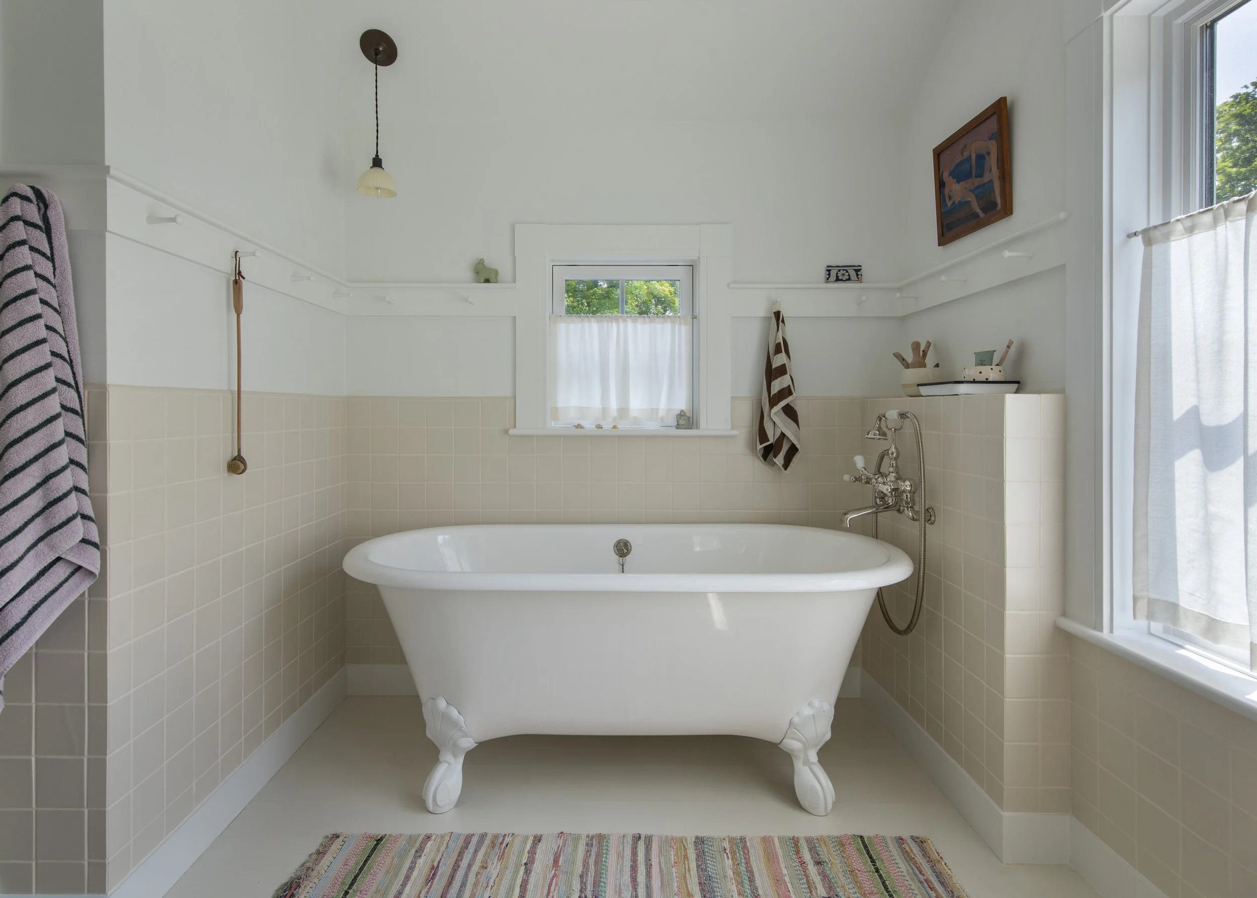 Photo of a vintage clawfoot bathtub in a white bathroom with a window, towels, artwork, and a colorful striped rug.