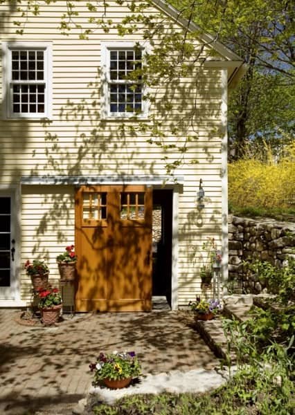 Front view of a white house with two upstairs windows and a wooden door below, with potted plants and flowers outside, and trees casting shadows on the house.