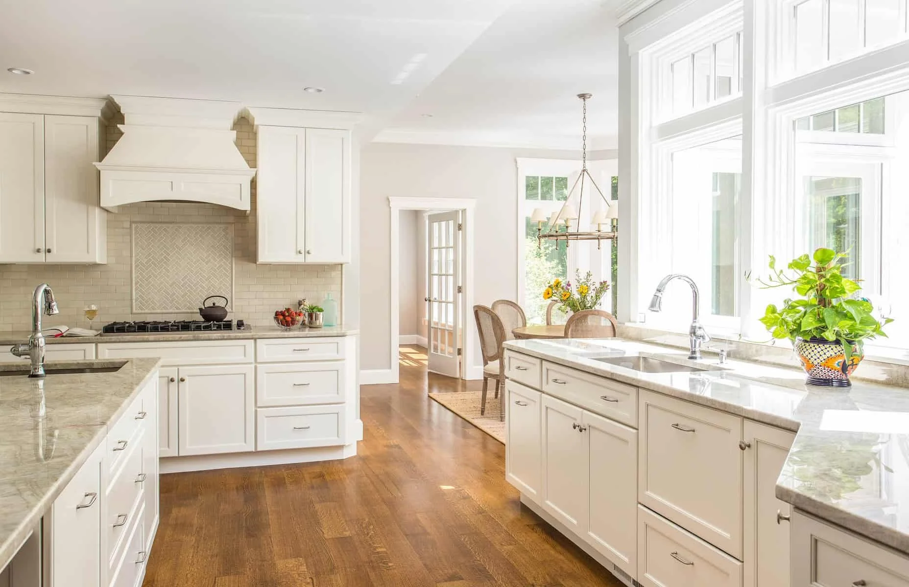 Bright kitchen with white cabinets, granite countertops, and large window over sink with a potted plant. Small dining area with a round table and chairs in the background.