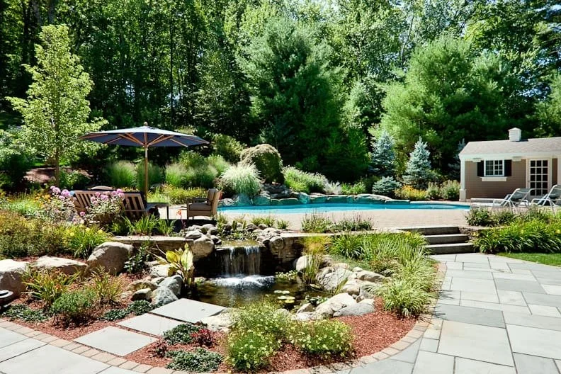 Backyard landscape featuring a small waterfall, a blue swimming pool, outdoor seating with an umbrella, a garden shed, and lush greenery with trees and shrubs.