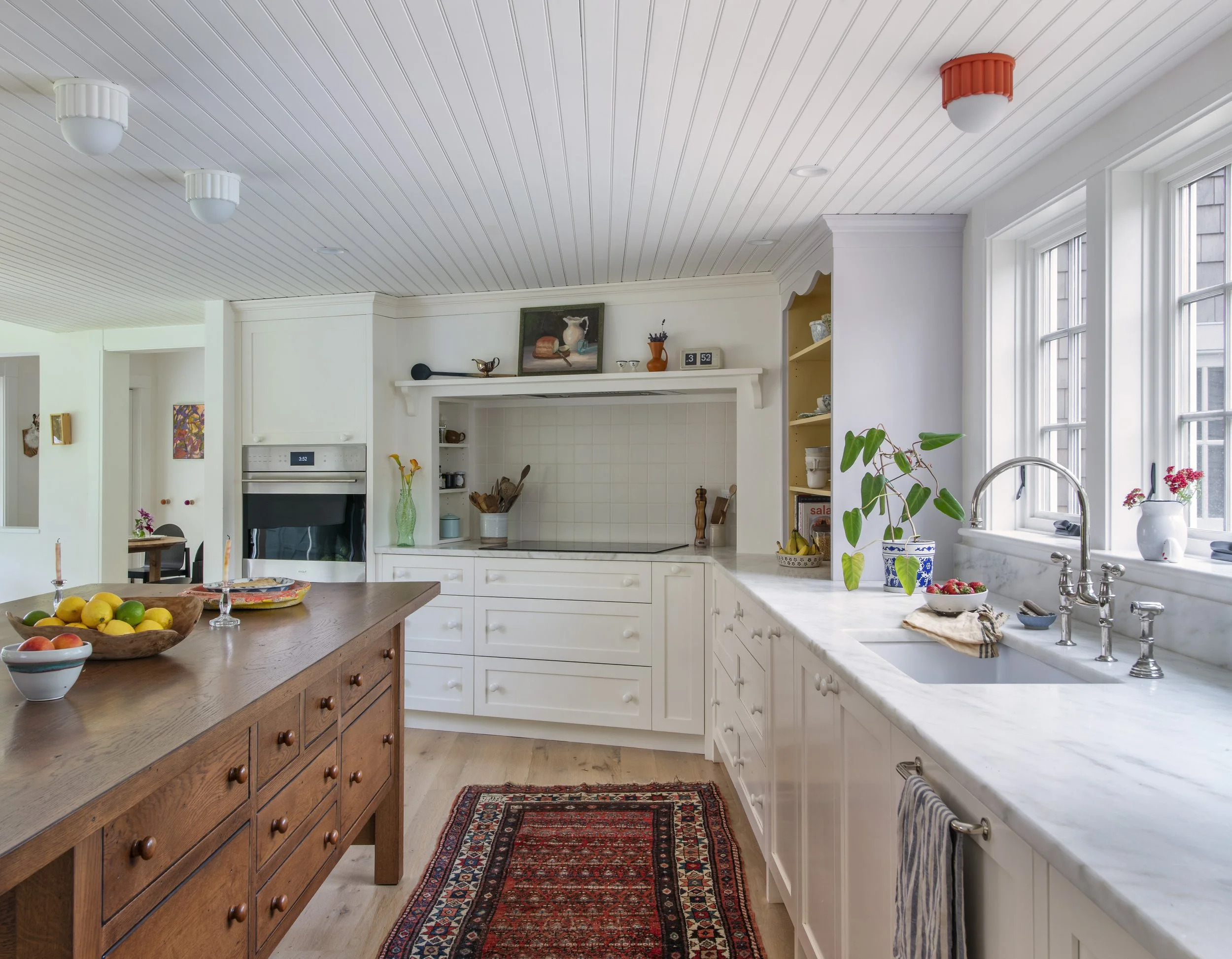 A kitchen with white cabinets, a marble countertop, a sink near large windows, and a wooden island with fruit and decor. Ceiling lights, a colorful rug, and decorative items on top of the cabinets are visible.