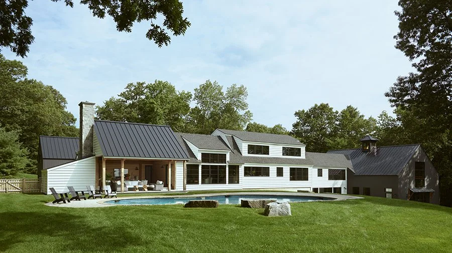 Modern house with white siding and black metal roof, large windows, a backyard with a swimming pool, patio furniture, and surrounded by green grass and trees.