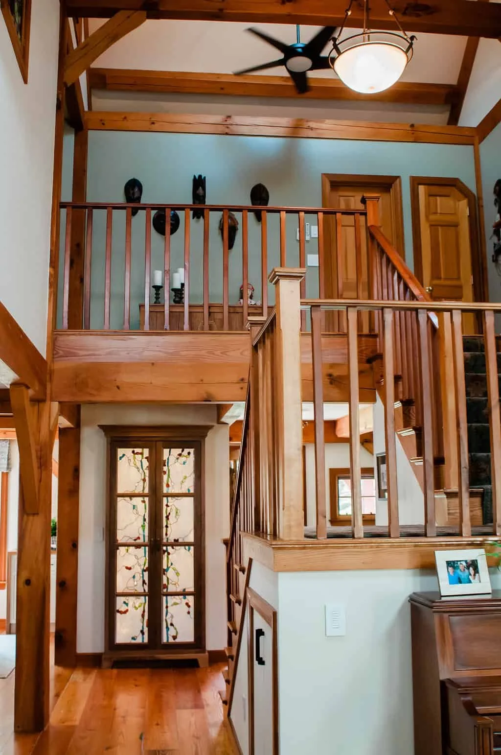 Interior view of a multi-level wooden staircase with a railing, ceiling fan, and decorative masks on the wall.
