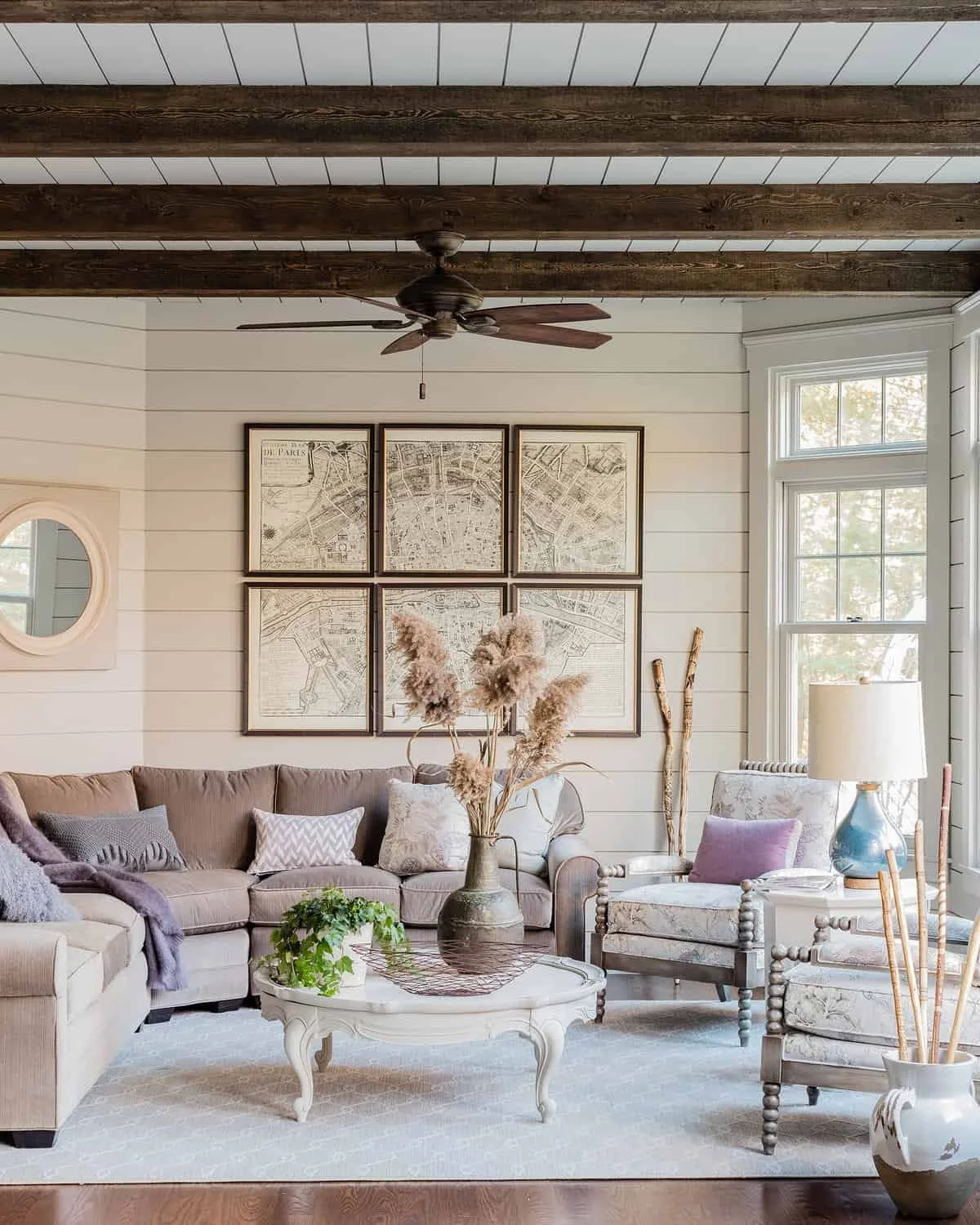 A living room with a beige sectional sofa, decorative pillows, a white coffee table with a vase of pampas grass, and a patterned armchair. The wall features framed vintage maps and a small round mirror, with a ceiling fan overhead and large window le