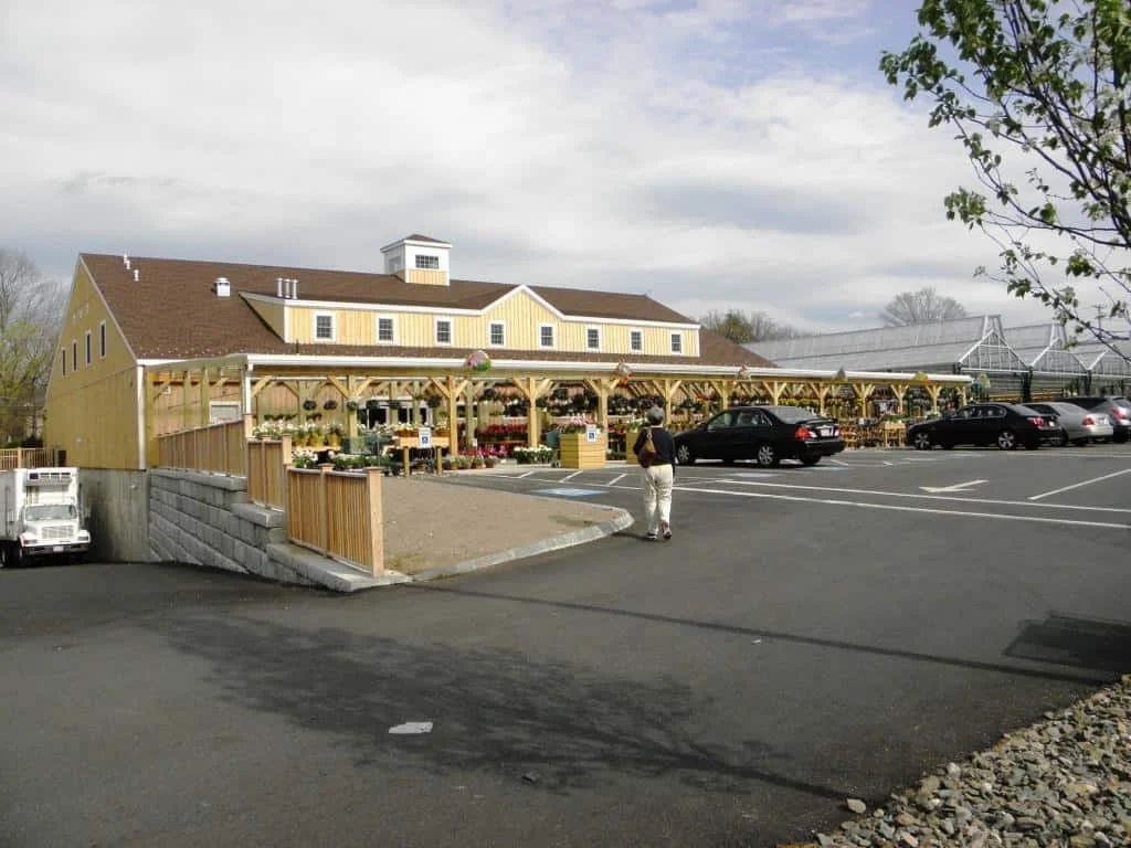 A yellow barn-style building with a garden center outdoor area, parking lot with several cars, and a person walking in the foreground.