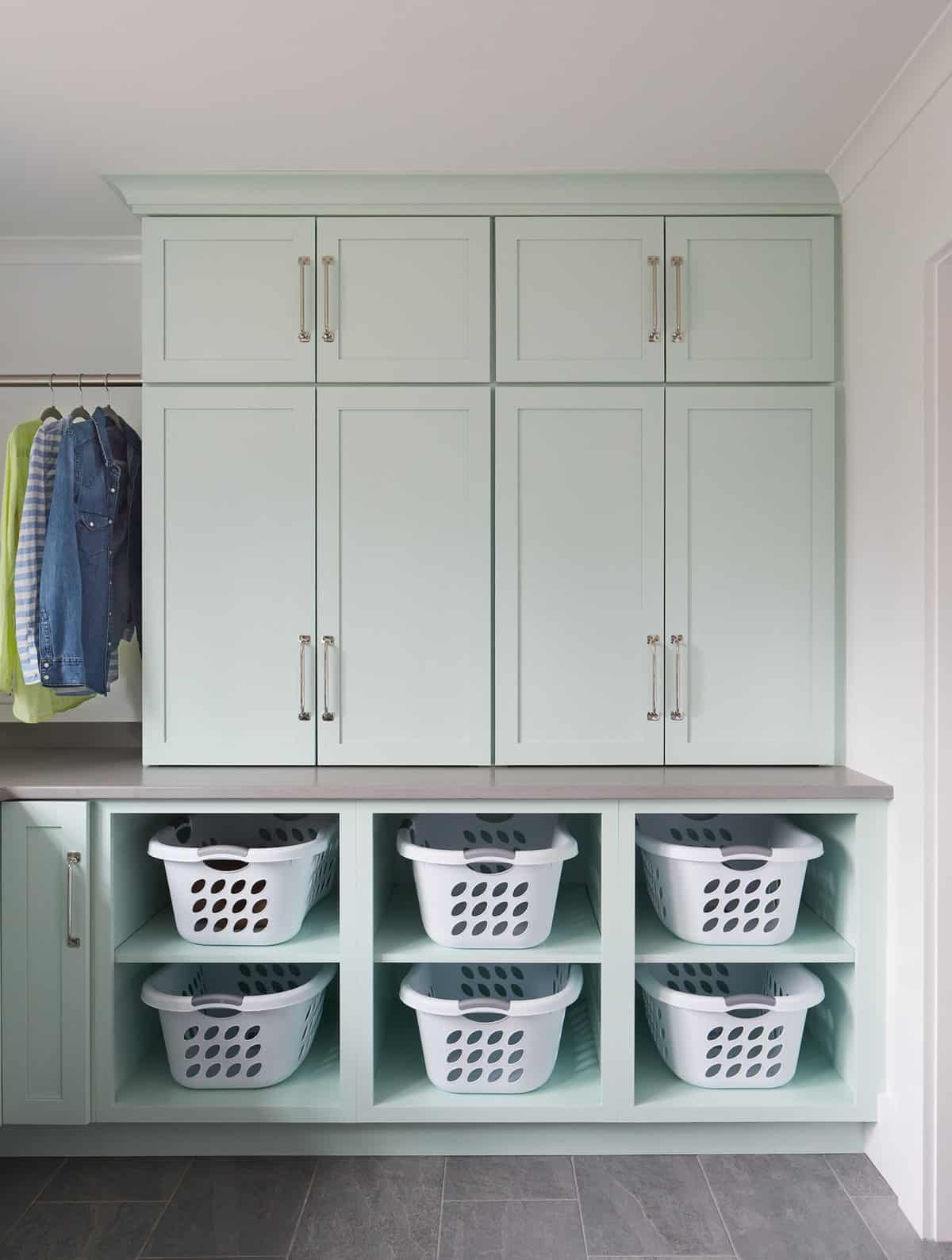 Light green built-in closet with three upper cabinets, two middle cabinets, and a lower section with eight cubbies holding white laundry baskets. To the left, a clothing rack with jackets and shirts hangs on a silver rod.