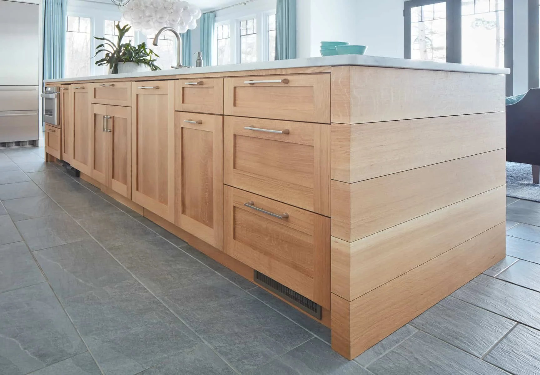 A modern kitchen island with wooden cabinetry, a marble countertop, and a built-in sink. There are additional cabinets above and around the island. Blue bowls are stacked on the countertop. Large windows with blue curtains let in natural light, and p