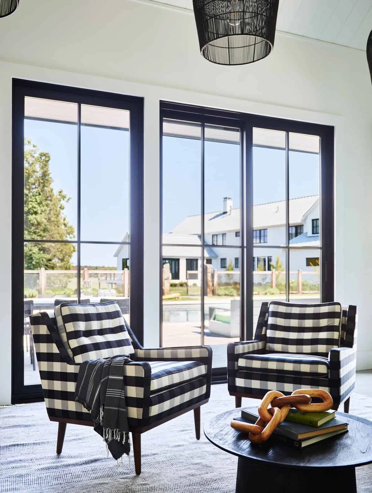 Living room with two black and white striped armchairs, a round black coffee table with books and a decorative chain sculpture, large window with black frames showing an outdoor scene with houses, trees, and a pool.