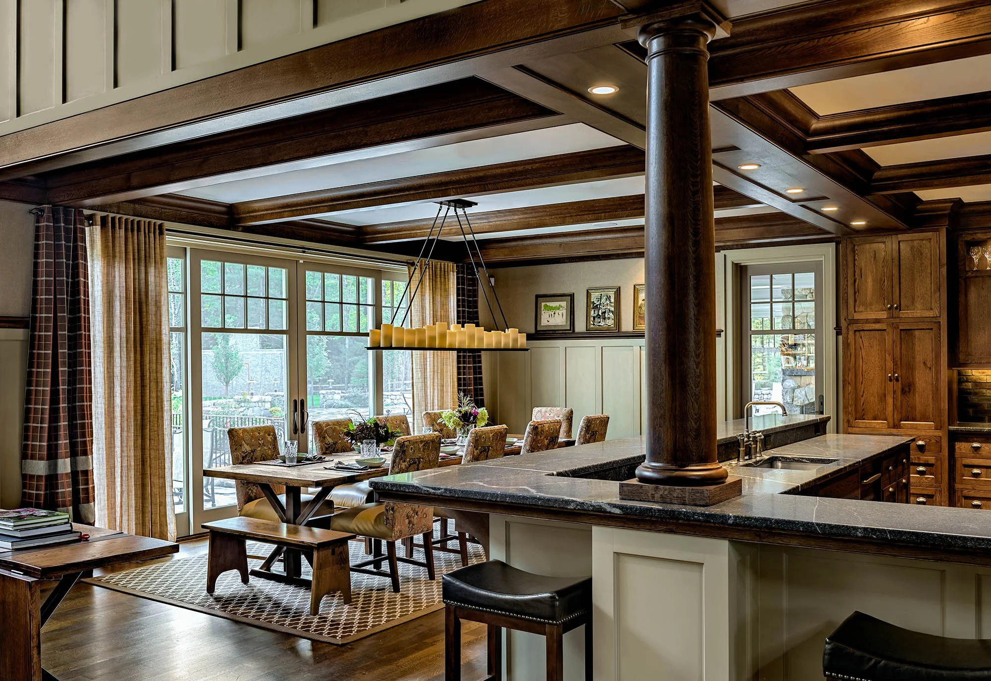 A cozy dining area with a large wooden dining table surrounded by floral upholstered chairs, set with plates and glasses, bathed in natural light from large windows with curtains, adjacent to a kitchen with wooden cabinetry, a granite countertop, and