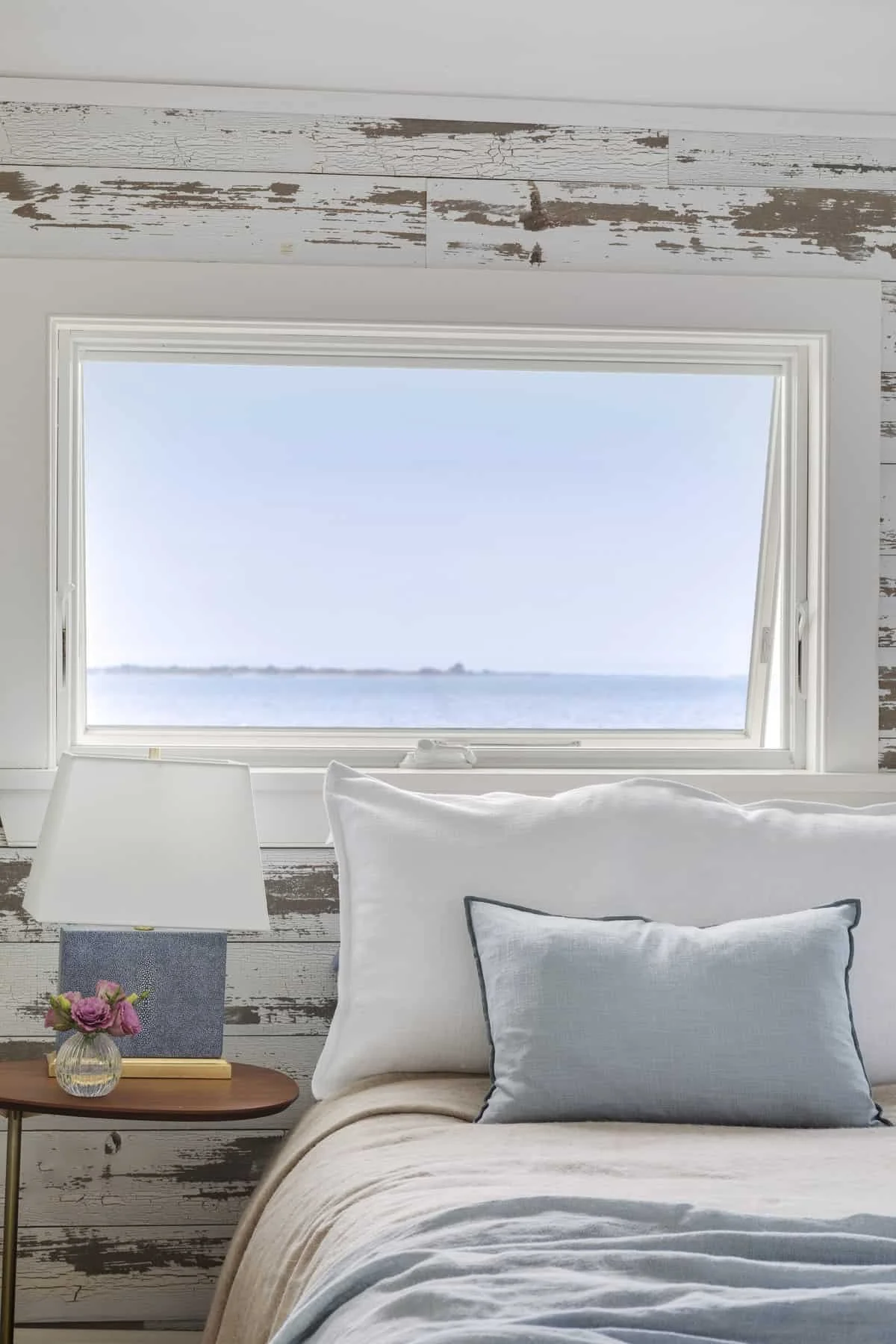 A bedroom with a large window offering a view of water and a distant shoreline. The bed has white and light blue pillows, and a beige blanket. Next to the bed, there is a small wooden nightstand with a flower vase and a lamp. The wall behind the bed 