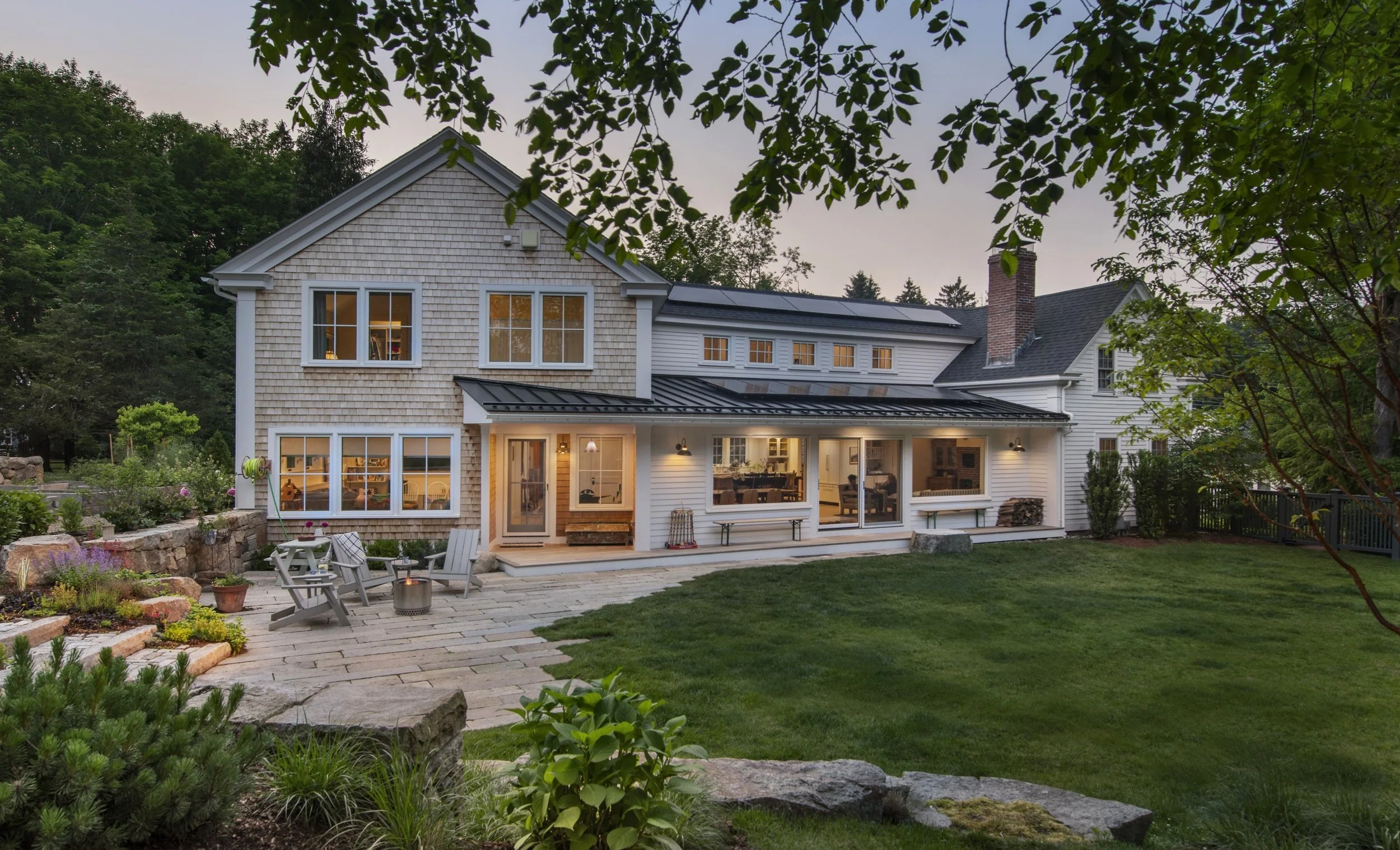 A two-story house with white siding and shingle exterior, large windows, and a black metal roof with solar panels, surrounded by lush green trees and a well-maintained backyard with outdoor seating and stone patio.