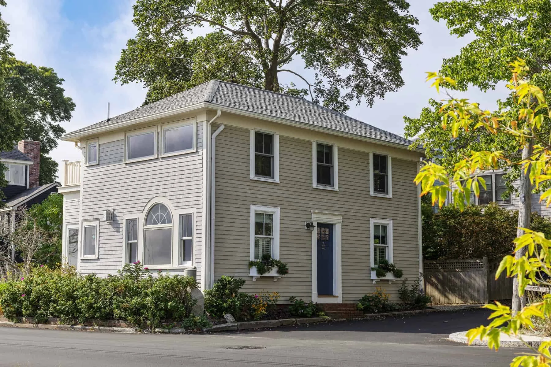 A two-story beige house with white trim, multiple windows, and a blue door, surrounded by green trees and landscaped bushes, with a street in front on a clear day.