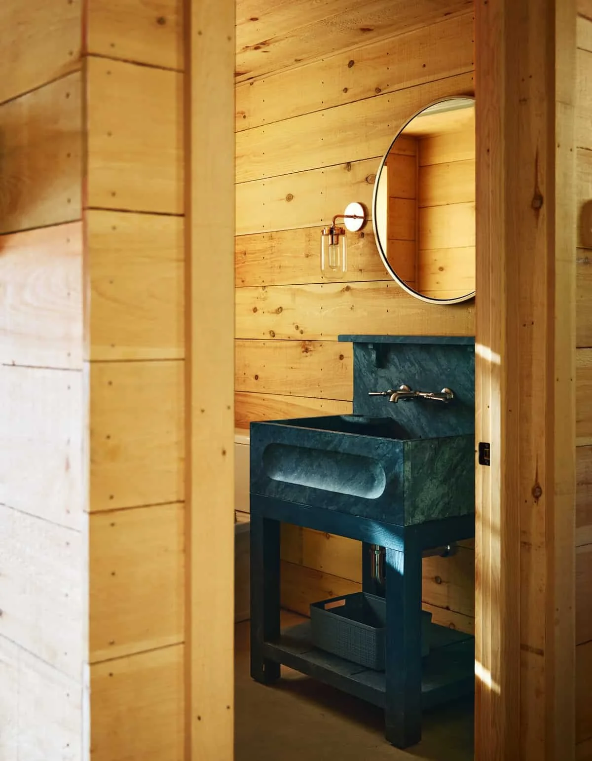 A rustic bathroom with wooden walls and ceiling, featuring a dark green marble sink on a black stand, a round mirror, and a wall-mounted light fixture.