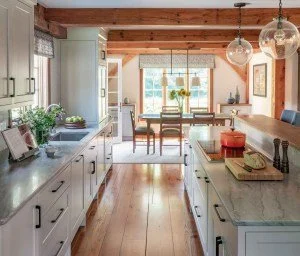 Bright kitchen with white cabinets, marble countertops, and large windows overlooking a patio. Wooden ceiling beams and pendant lights above the island.