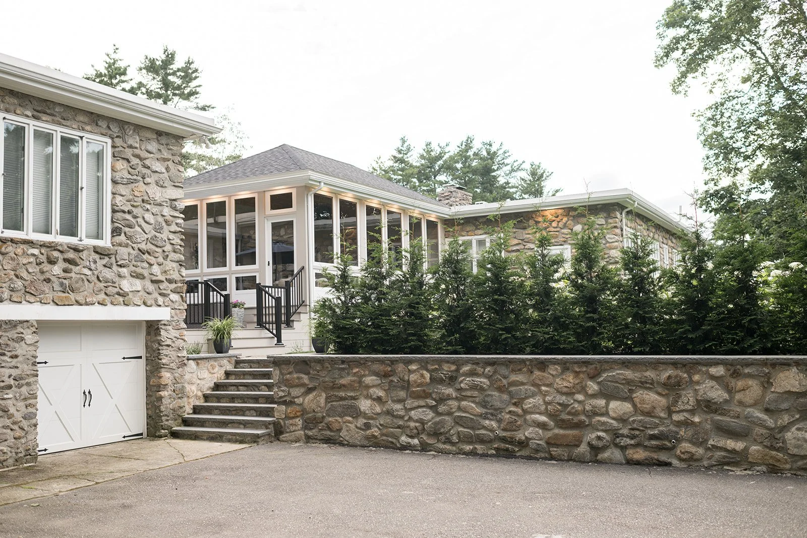 A house with a stone exterior, a white garage door, and a porch with stairs, surrounded by trees and greenery.