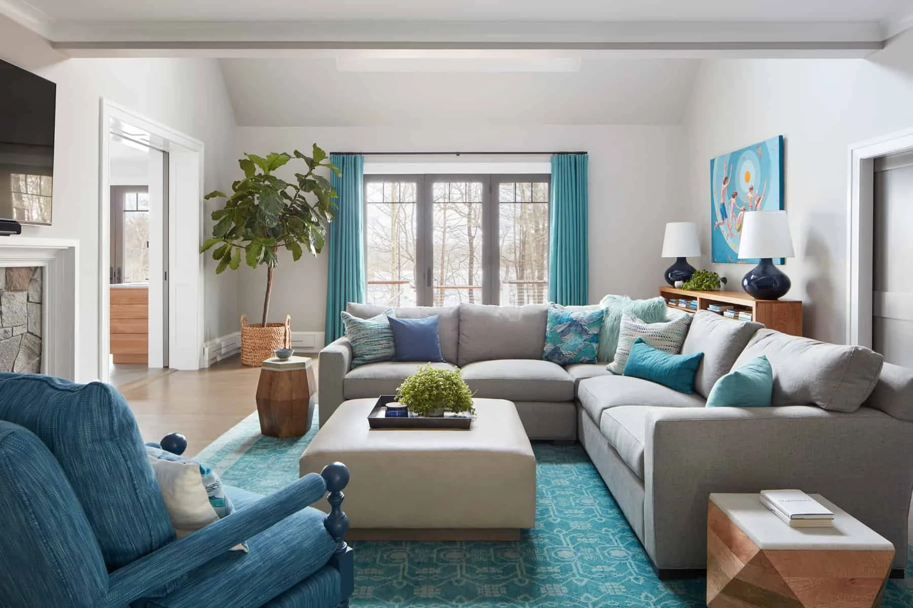 Bright living room with large sectional sofa, blue armchair, and colorful pillows, large window with blue curtains, potted plant, abstract wall art, and wooden side tables with lamps.