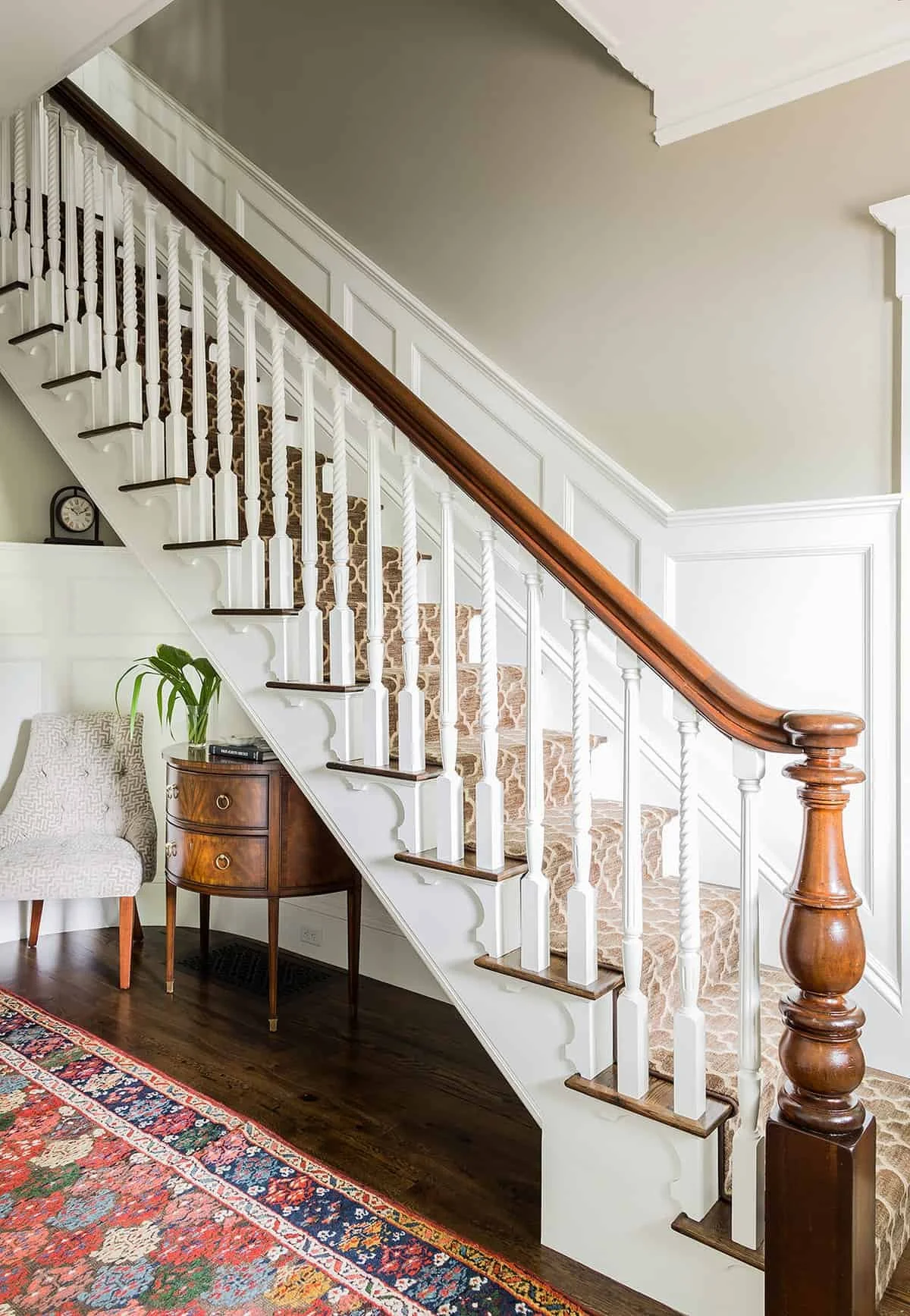 Interior view of a staircase with white spindles, a wooden handrail, and a patterned carpet on the stairs. In the foreground, there is a wooden side table with decorative items, a potted plant, and a beige armchair with a patterned cushion. A colorfu
