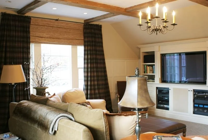 Living room with beige and cream sofas, a large window with plaid curtains, a white built-in entertainment center with a TV, a chandelier, and a table lamp.